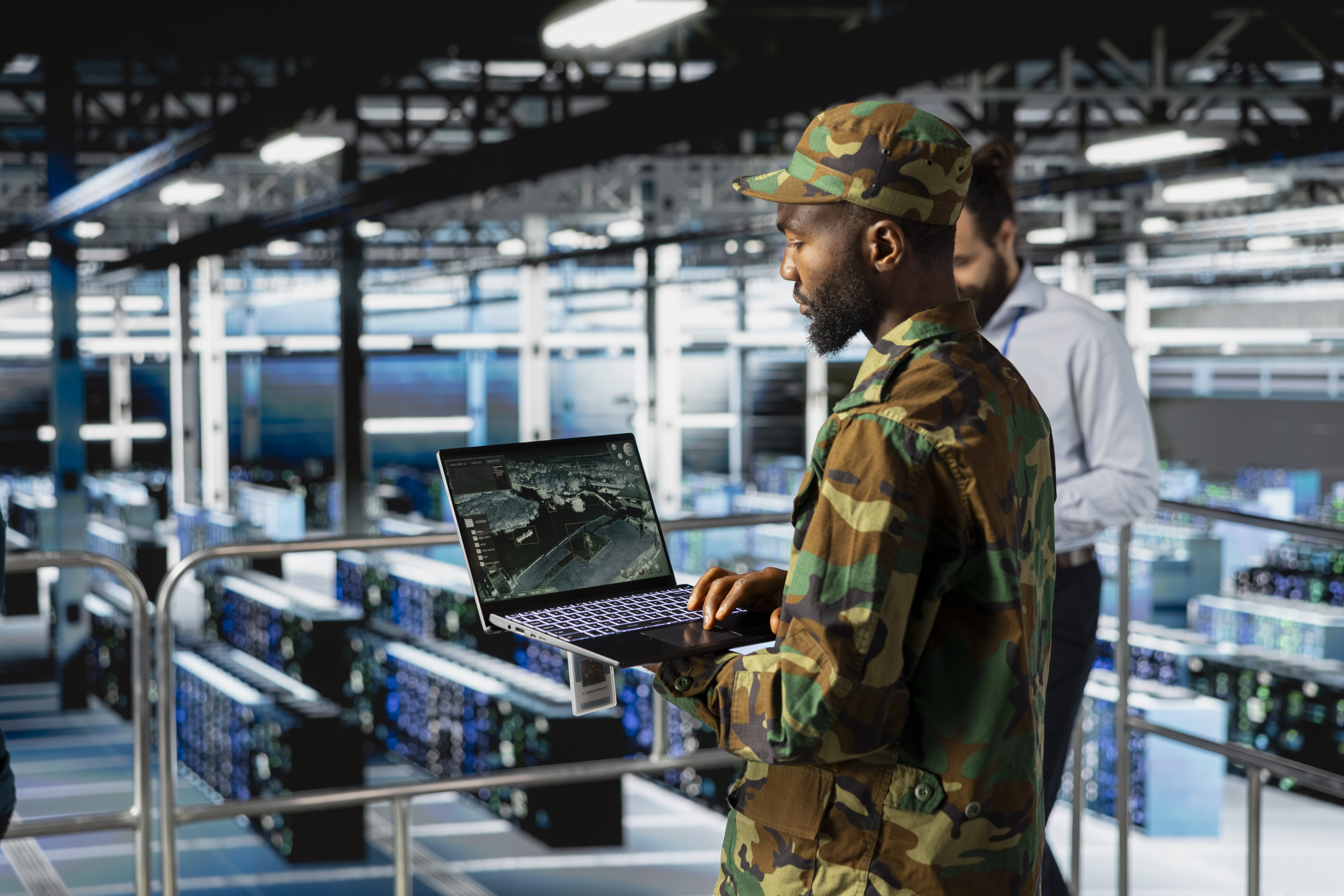 Pessoa em uniforme militar utilizando um laptop em ambiente tecnológico com servidores ao fundo.